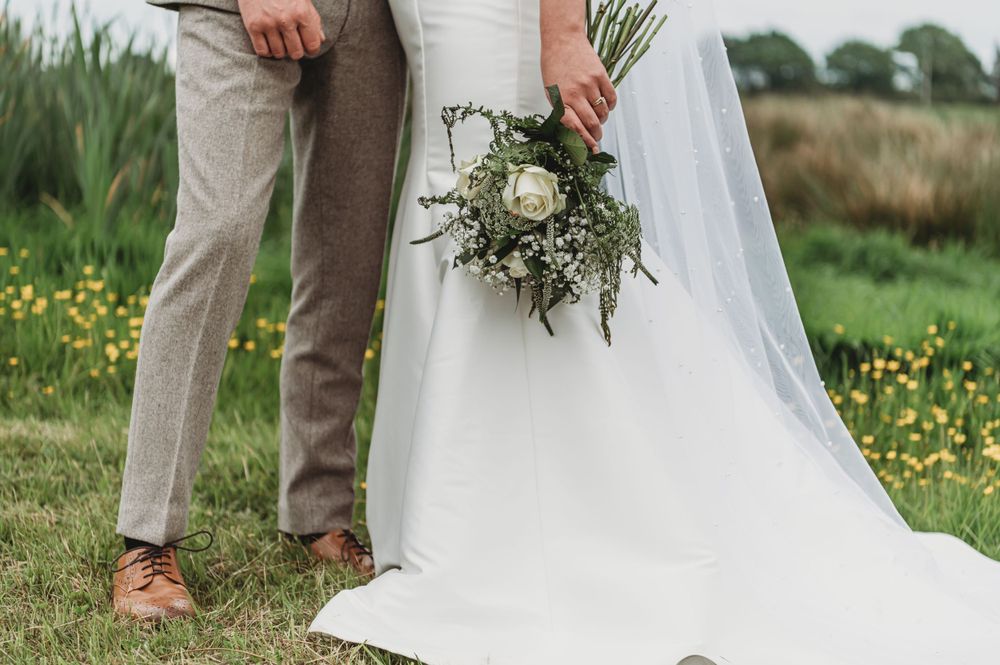 White wedding dress and bouquet detail shot against a green meadow with wildflowers.