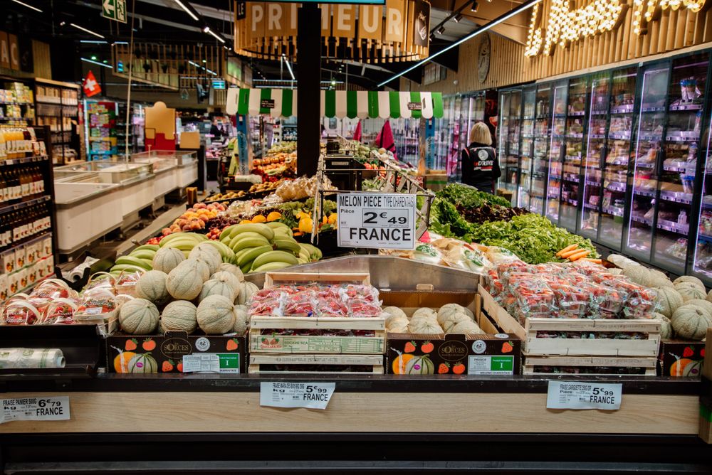 A colorful supermarket fruit and vegetable section with melons, bananas, and French-labeled produce on display.