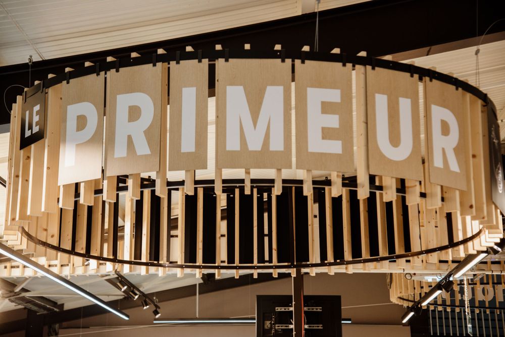 A decorative circular wooden sign reading Le Primeur hanging from the ceiling of a modern food market.