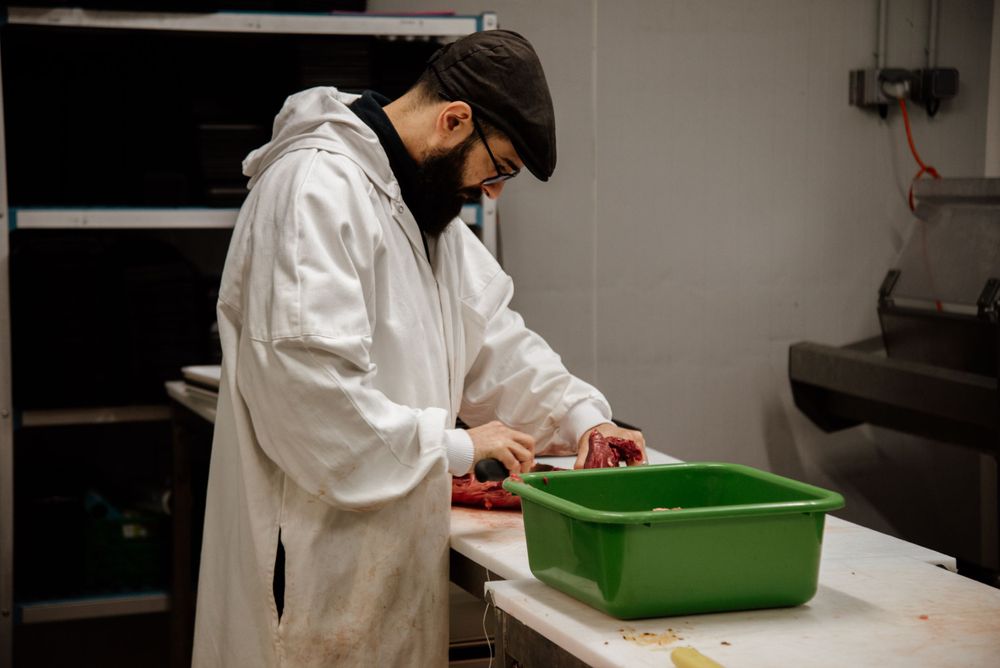 Butcher in white coat and cap preparing fresh meat on a clean work table in a professional kitchen.