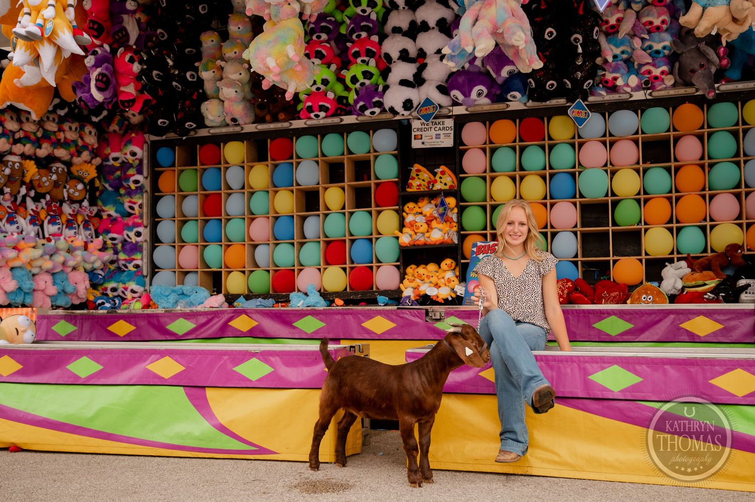 A show goat stands near a vibrant carnival game booth filled with colorful stuffed animal prizes.