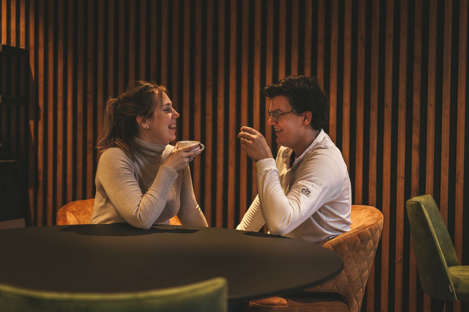 People engaged in conversation at a dimly lit restaurant with wooden panel walls in the background.
