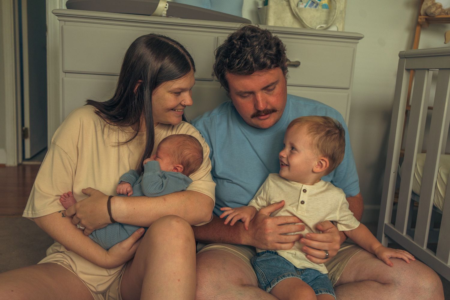 Young family with newborn and toddler sitting together in a nursery room.