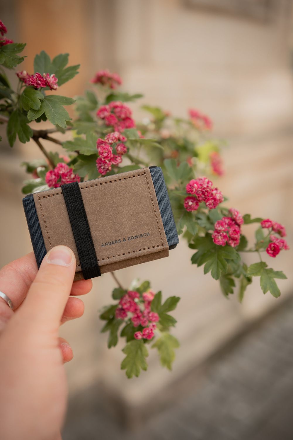 Hand holding slim leather wallet against blooming pink flowers and green leaves.