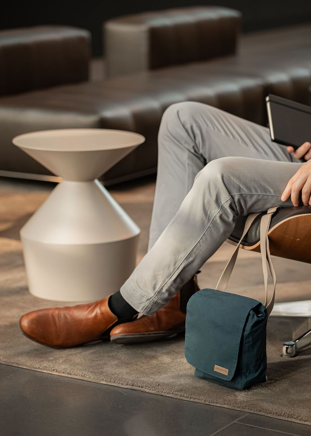 Modern interior scene showing brown leather shoes, gray pants and a teal messenger bag against wooden flooring.