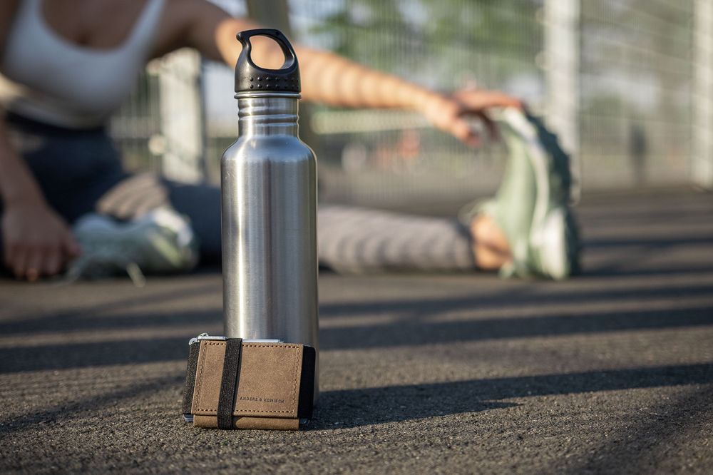 Metal water bottle with wooden holder sitting on dark asphalt surface.