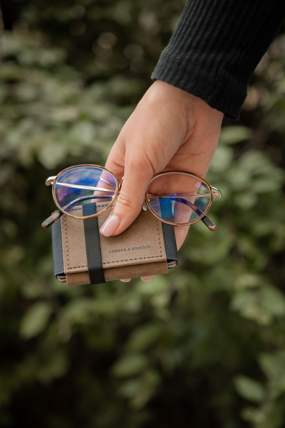 Hand holding eyeglasses on brown leather wallet against blurred green foliage background.