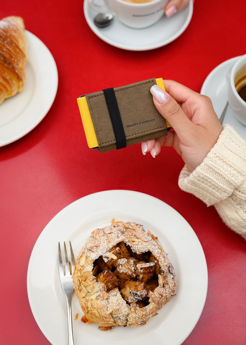 Leather wallet held over breakfast scene with pastries and coffee on red table.