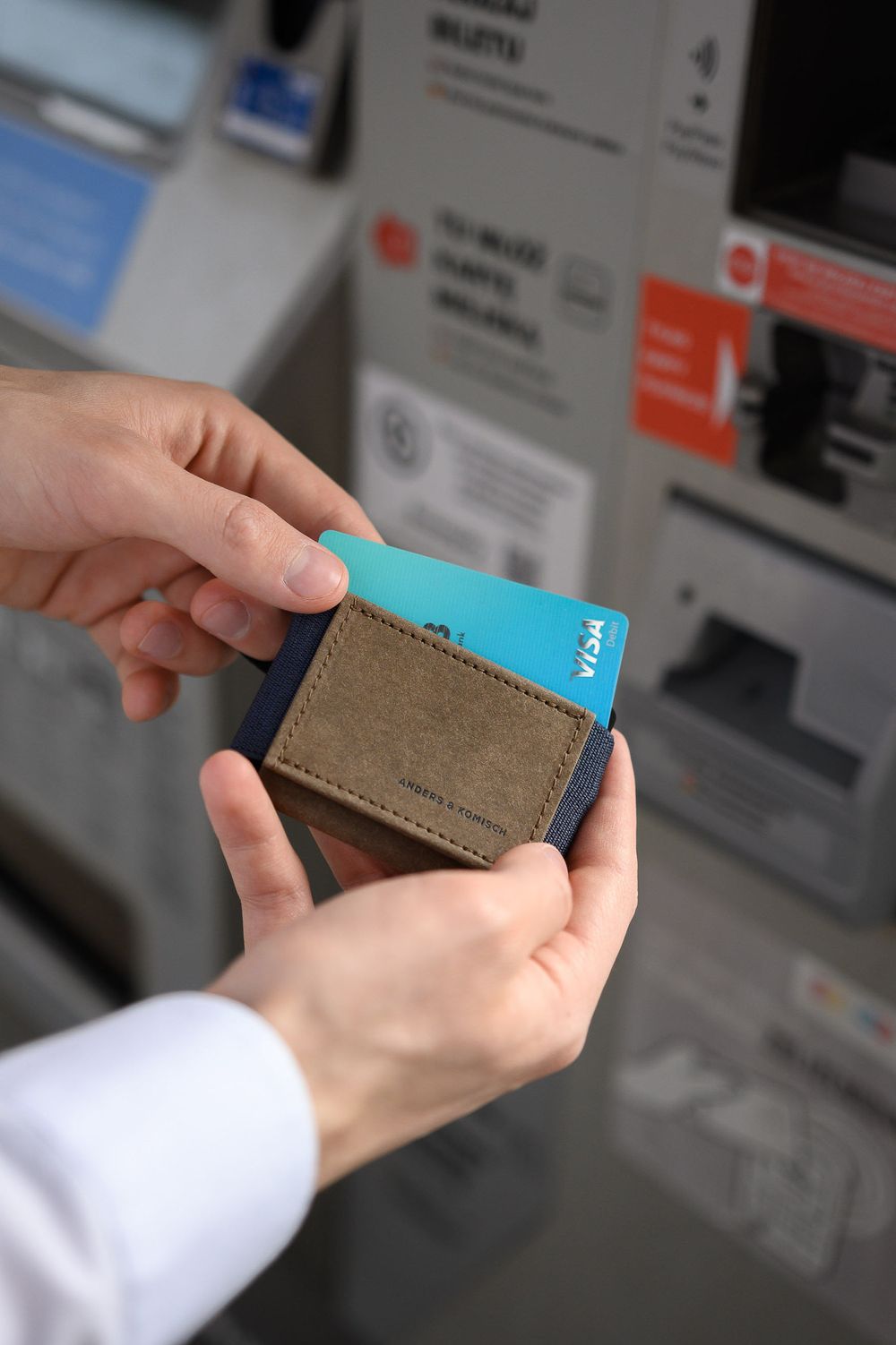 Hands inserting blue card into slim brown leather wallet at ATM machine.