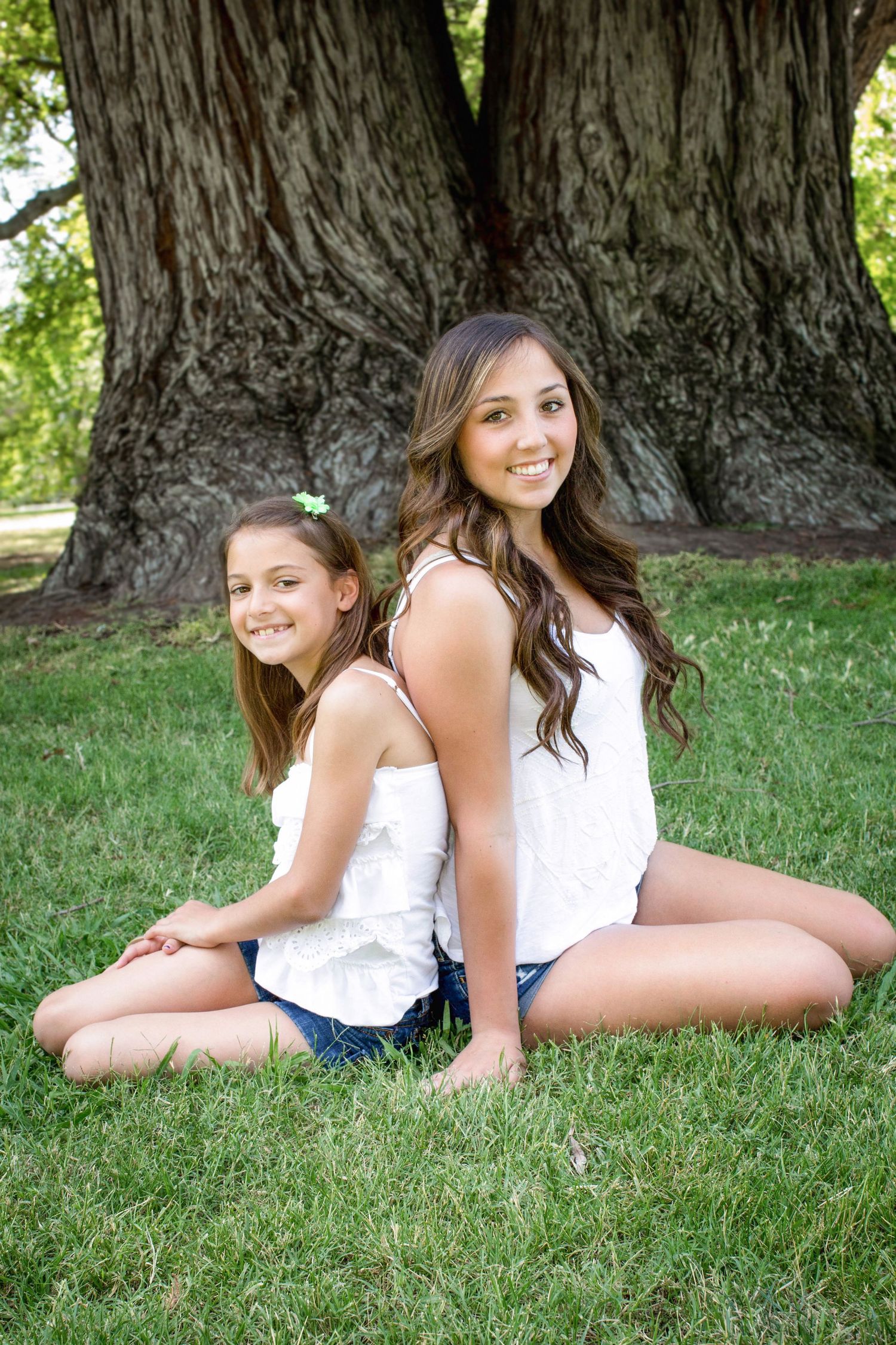 Two people sitting back-to-back against large tree trunk on grass.
