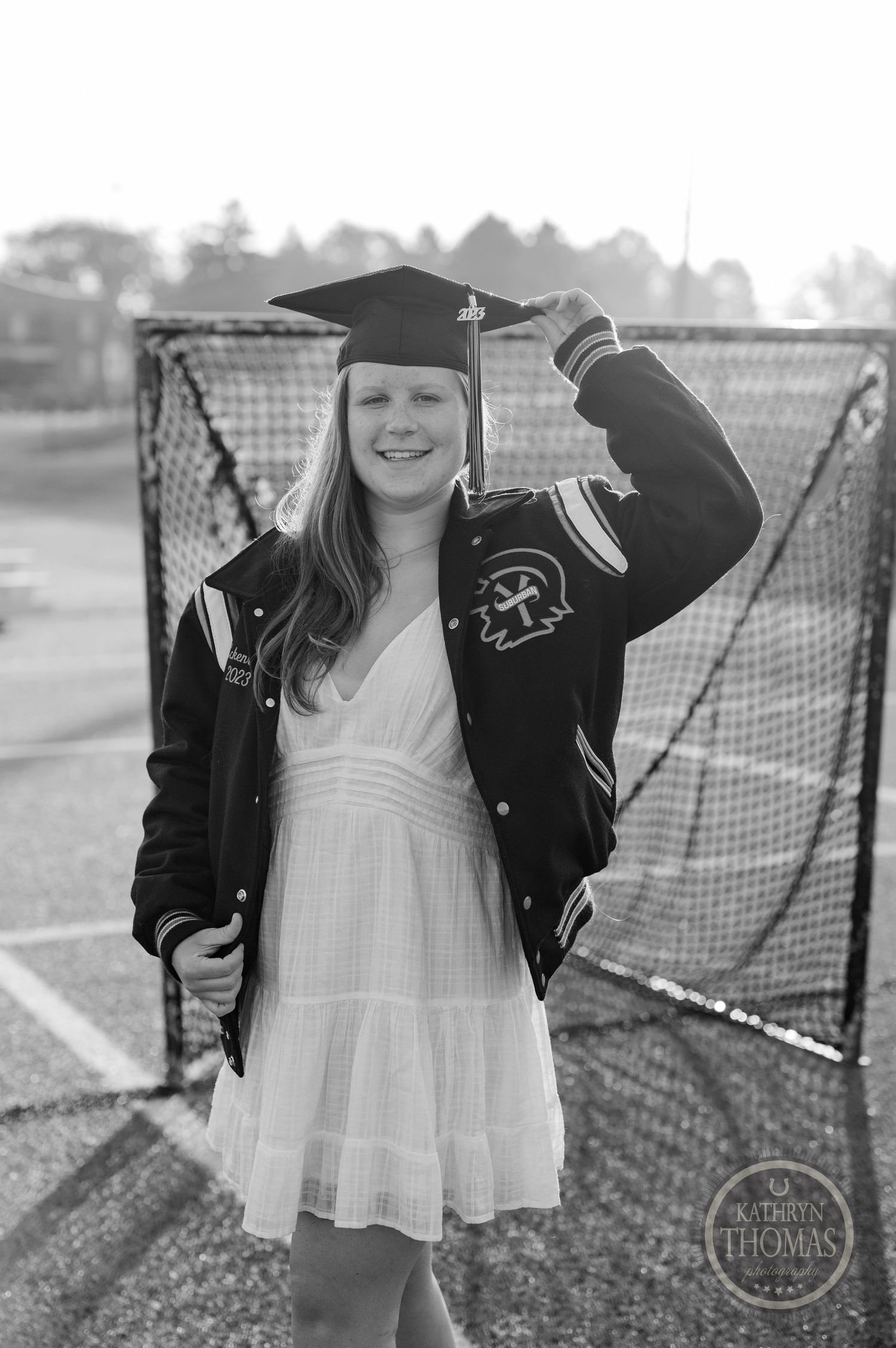 Graduate in varsity jacket and cap poses on athletic field with lacrosse goal in background.