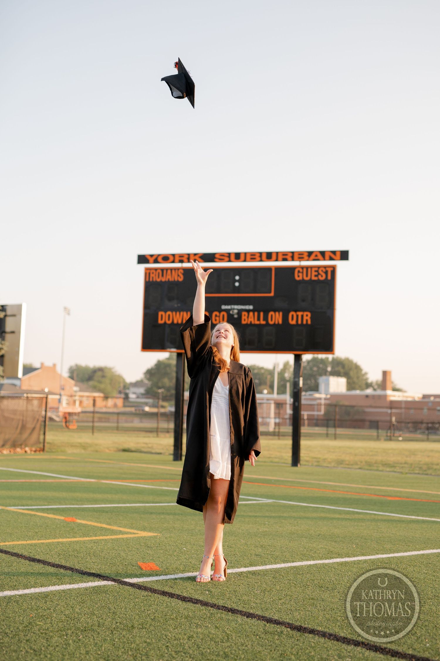 Graduate in white dress and black gown tosses cap in celebration on athletic field with scoreboard in background.