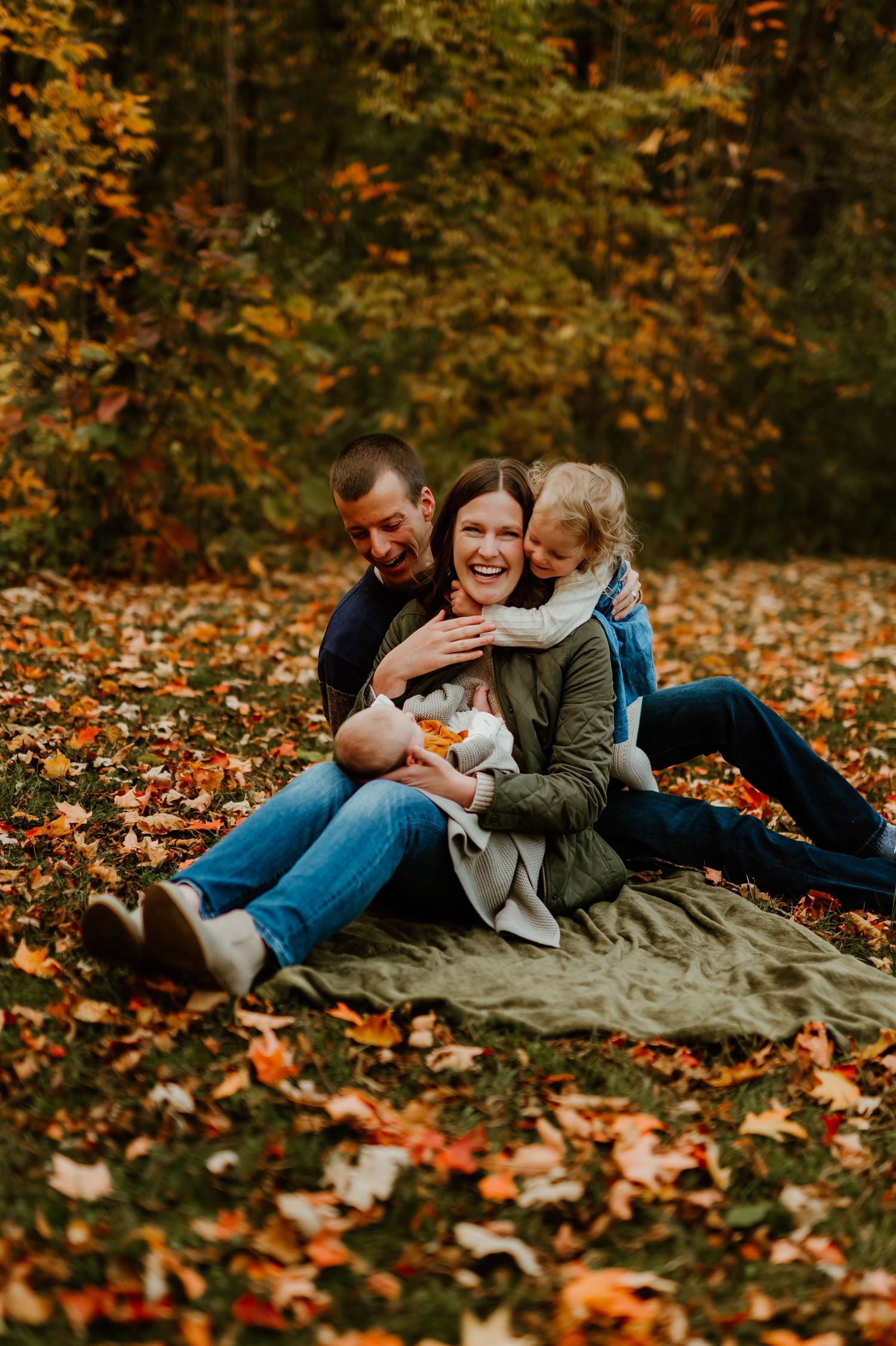 Family of four laughing together on a blanket surrounded by colorful autumn leaves in a forest setting.