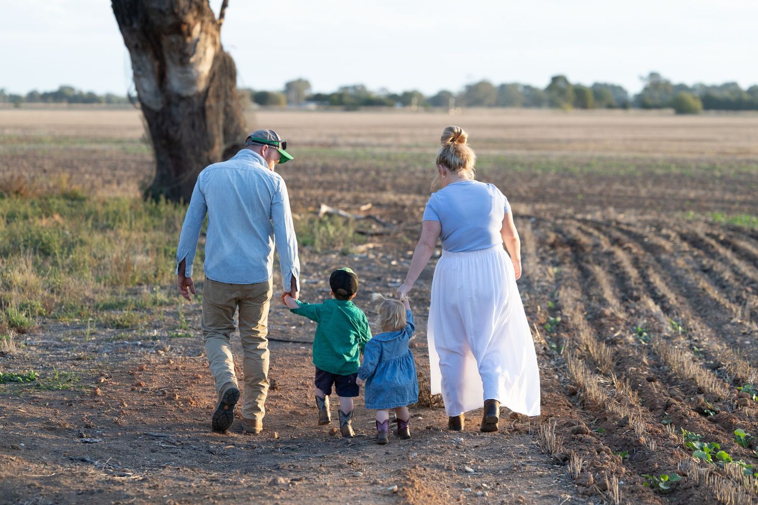 Family of three walking hand-in-hand along a rural dirt path beside a tree in an open farm field.