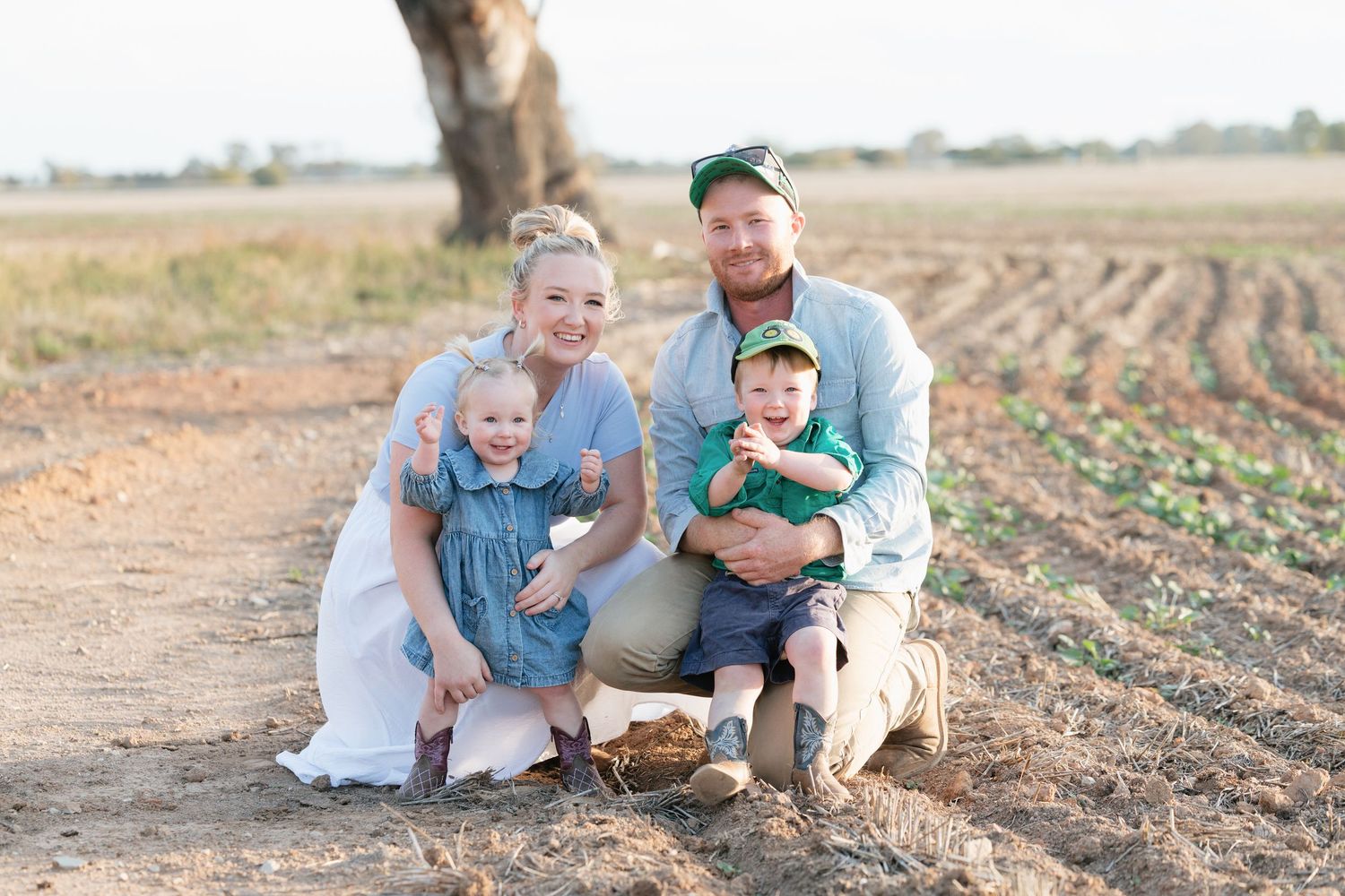 Family of four smiling together on a farm dirt path, parents with two young children on a sunny day.