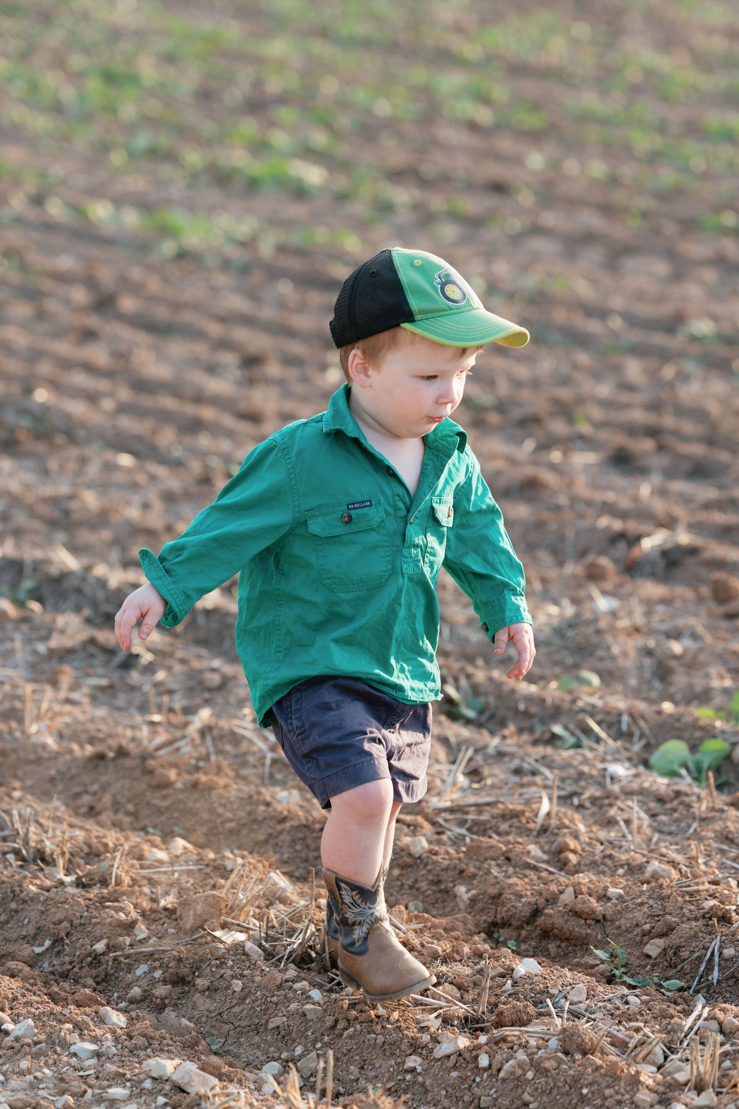 Young boy in green shirt and cap runs through a freshly plowed farm field wearing muddy boots.