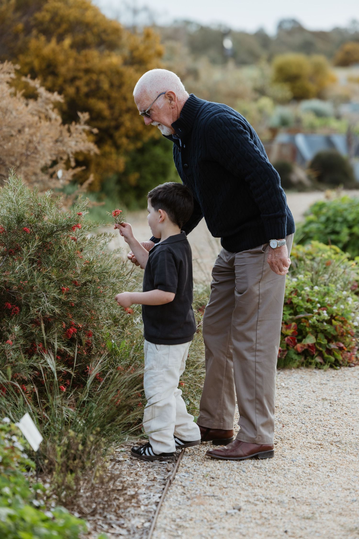 Two people explore a garden path together while looking at plants and flowers in an outdoor setting.