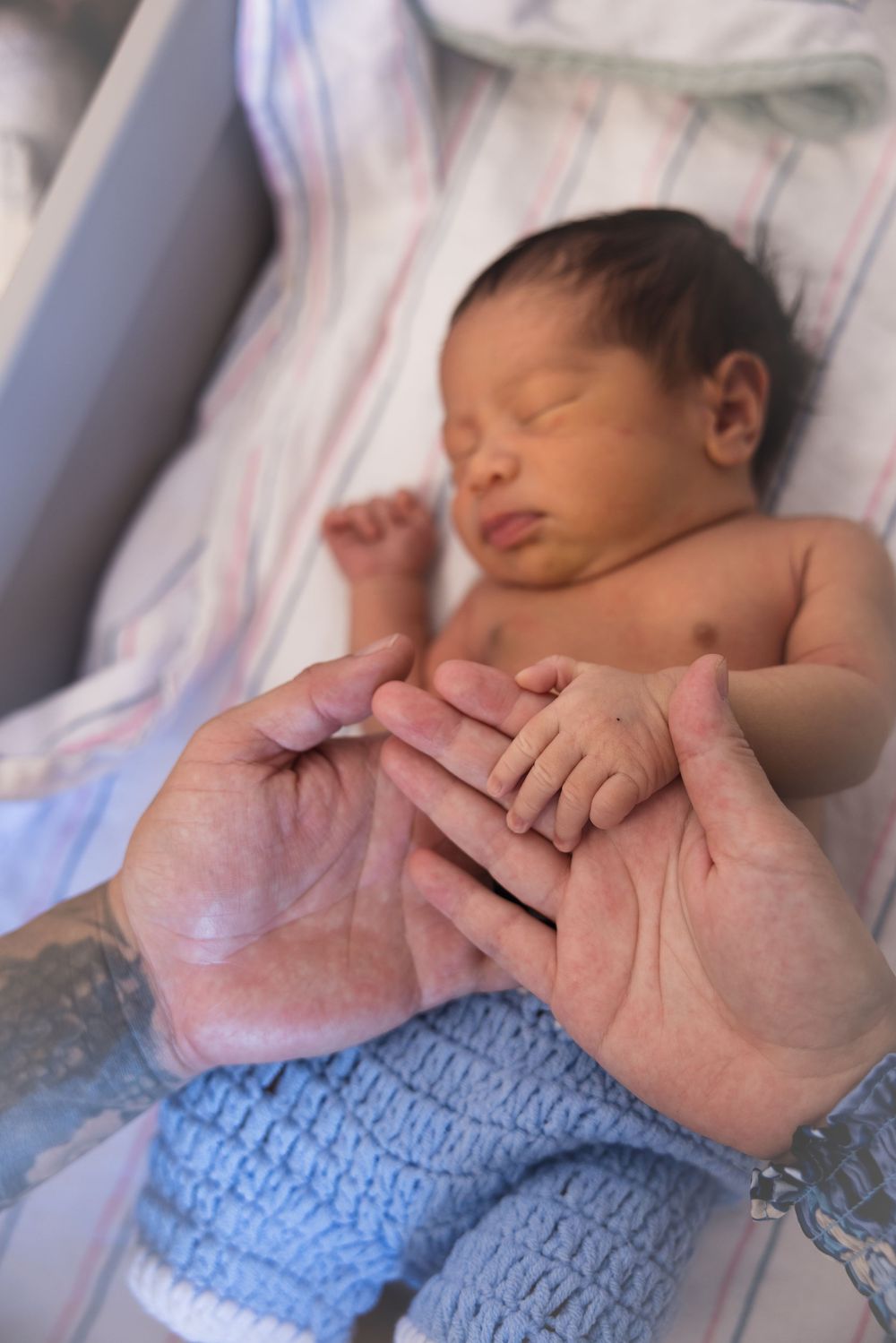 Close-up series of a sleeping newborn baby wearing a blue knit outfit while tiny hands are being held by adult hands.