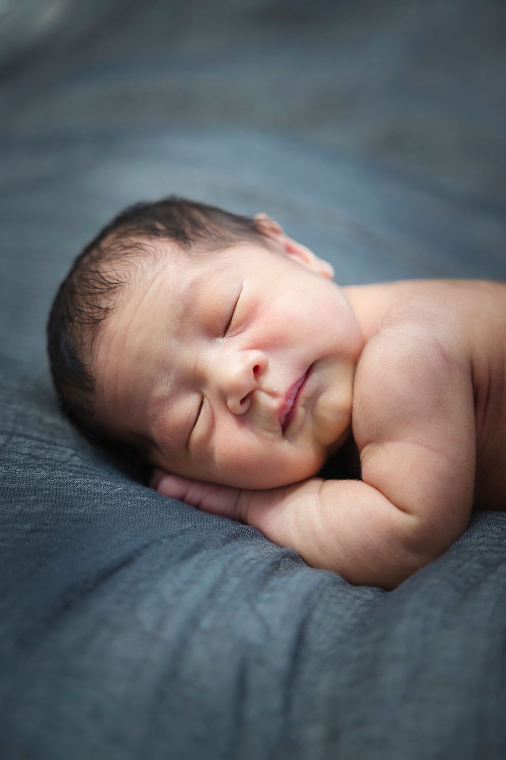 A peaceful sleeping baby rests on soft gray fabric with gentle lighting creating a serene newborn portrait.