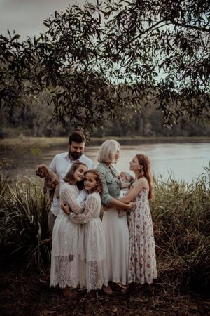 A family wearing white and floral dresses stands together by a peaceful lake at sunset under overhanging tree branches.