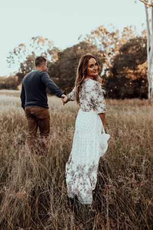 A couple holding hands walks through tall grass in a dreamy autumn field at sunset.