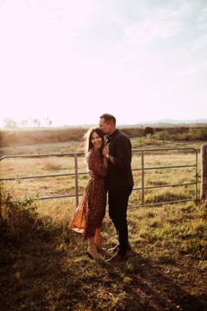 A couple shares a tender moment at sunset by a rustic fence in a golden countryside field.