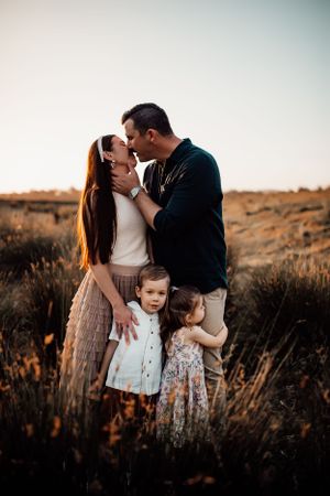 A family embraces in a field during golden hour as the sun sets over dried grass and brush.