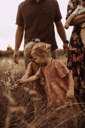 A child in a coral dress explores a grassy field at sunset while family members stand nearby.