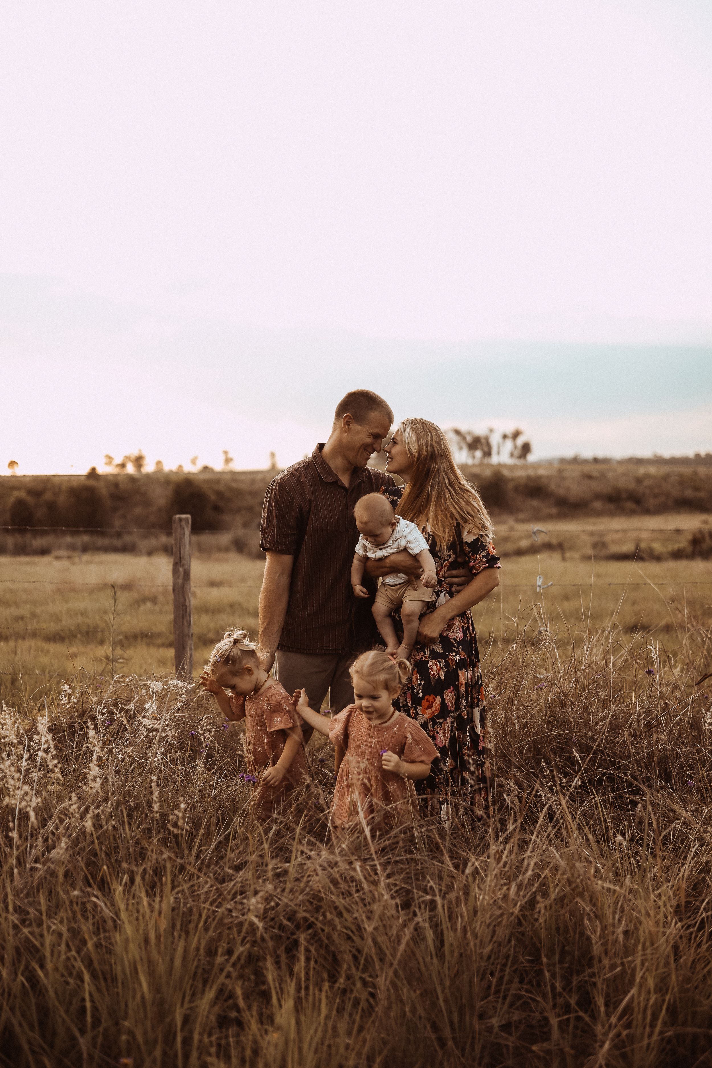 A family embraces together in a golden field at sunset while three young children play in the tall grass.
