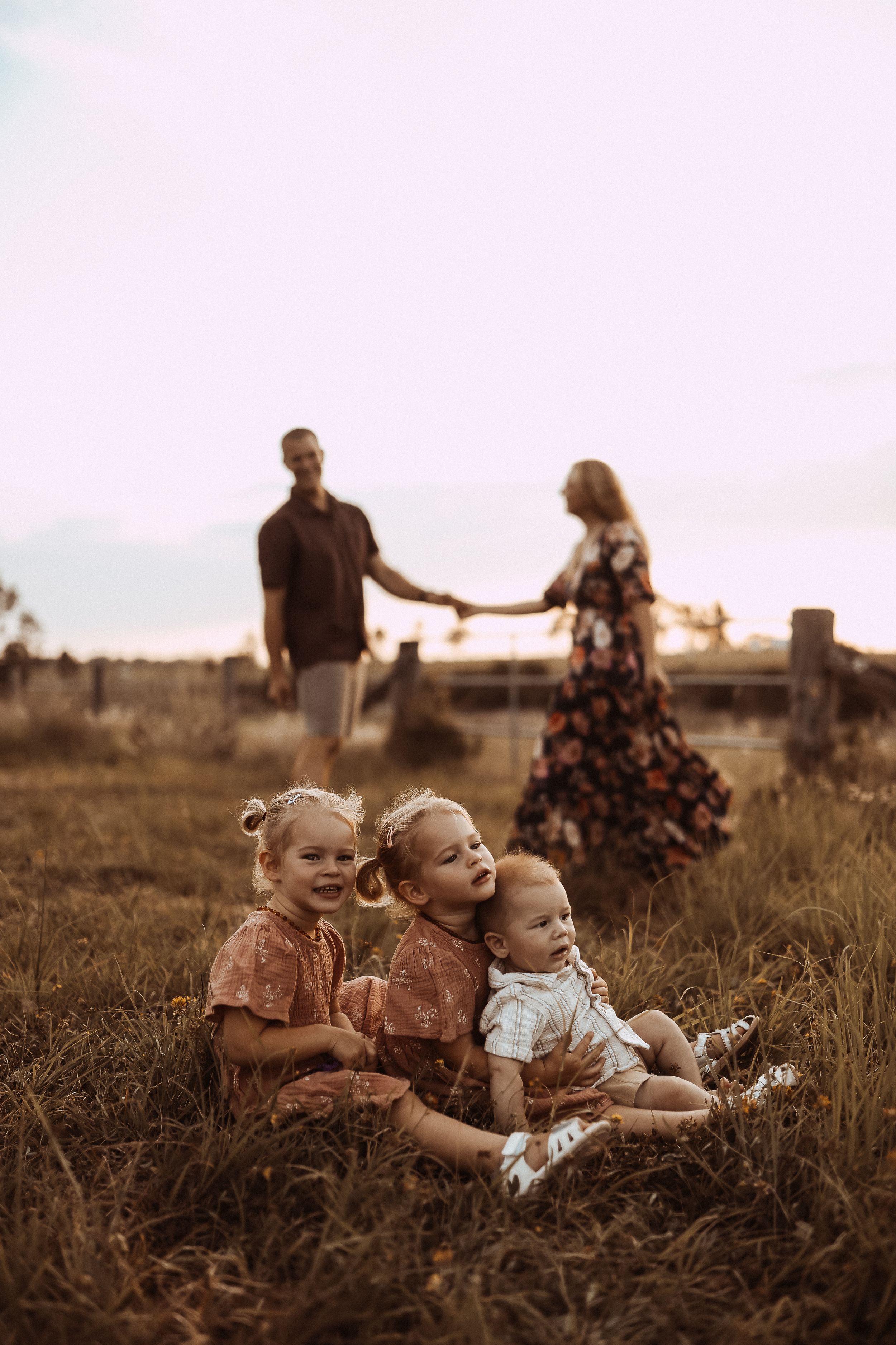Three children sit together in a grassy field at sunset while their parents hold hands in the background.