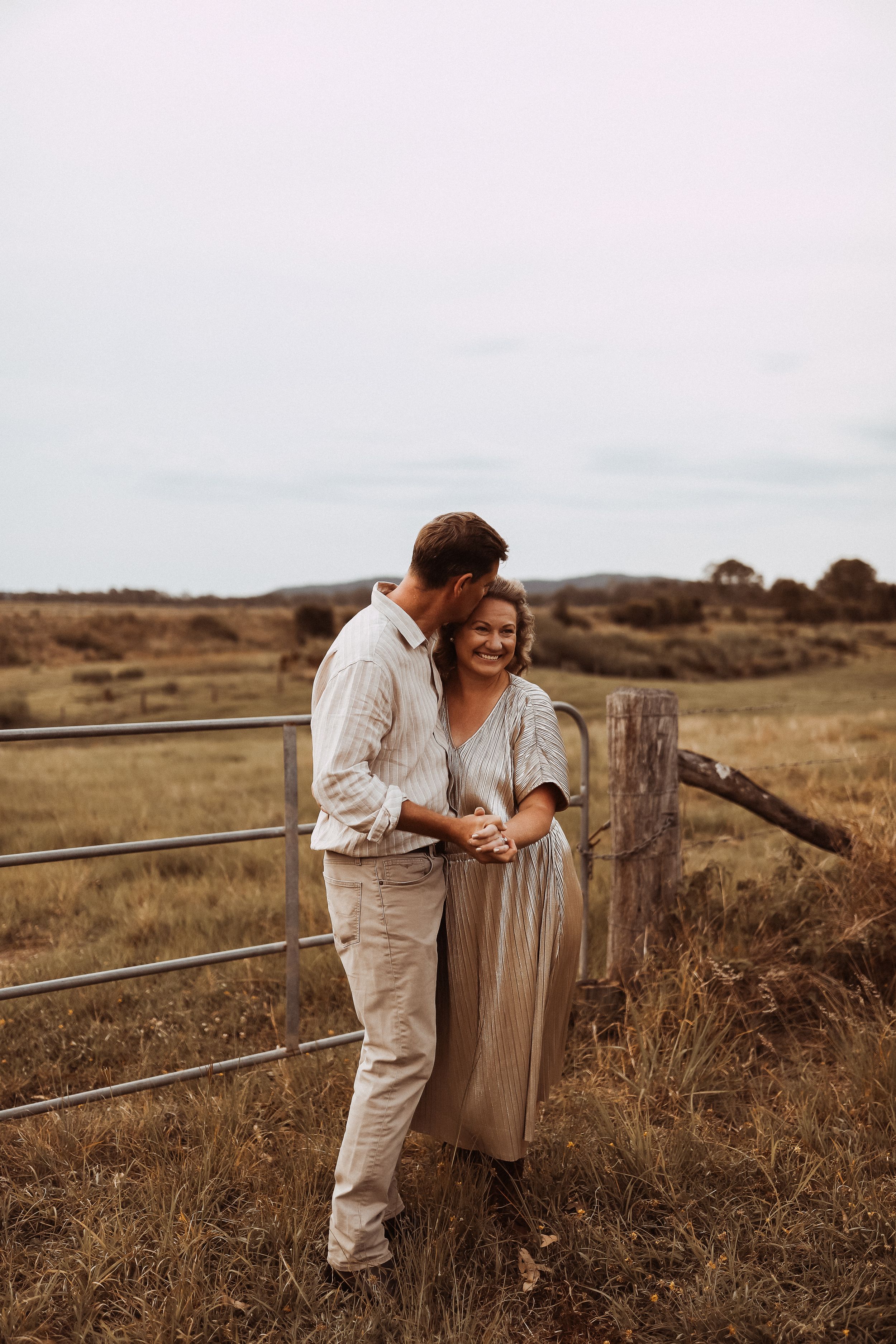 A romantic countryside scene with a couple embracing by a rustic fence during golden hour in a dry grassy field.