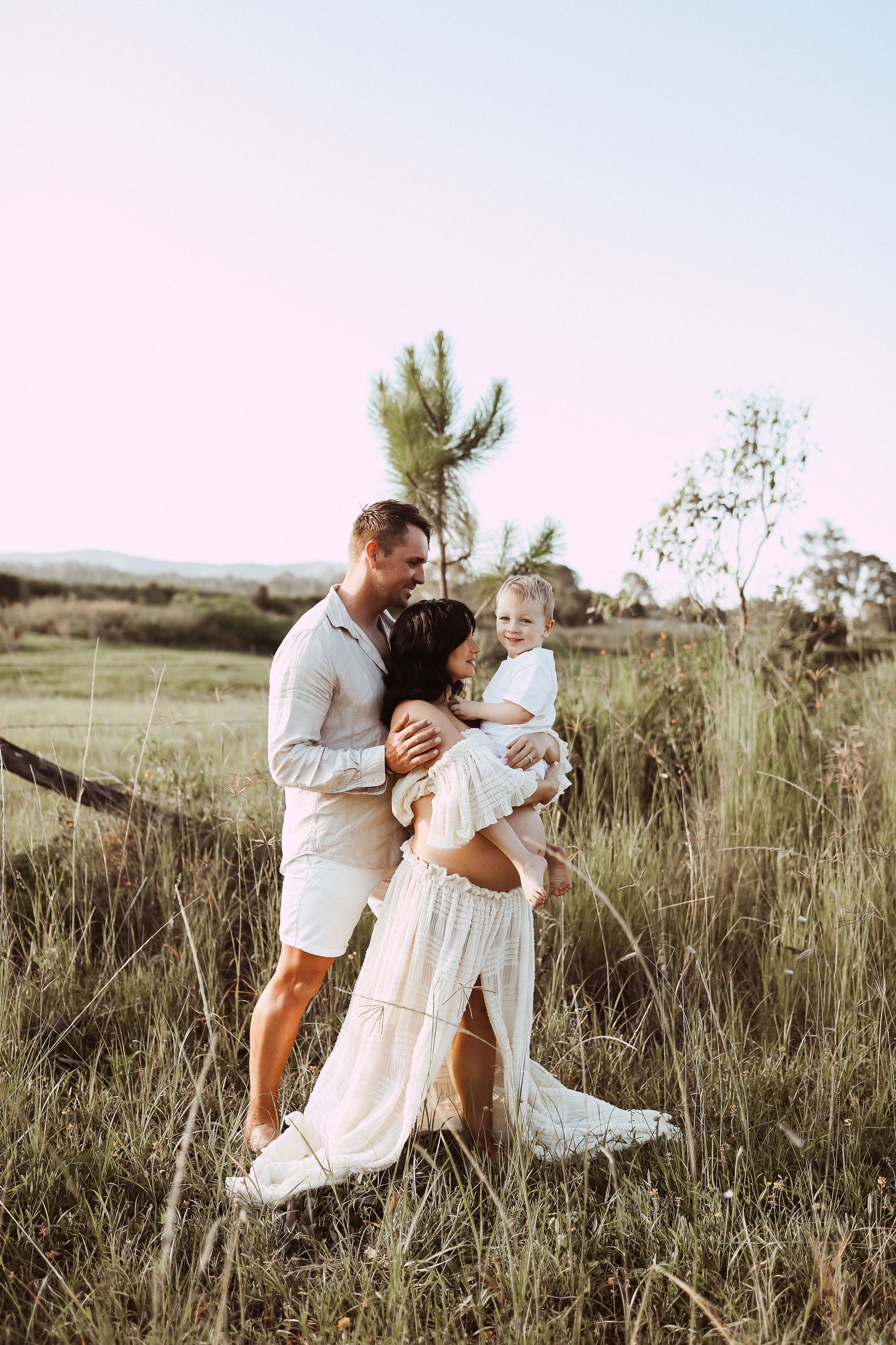 Couple embraces in sunlit field with tall grass and pine trees in the background.