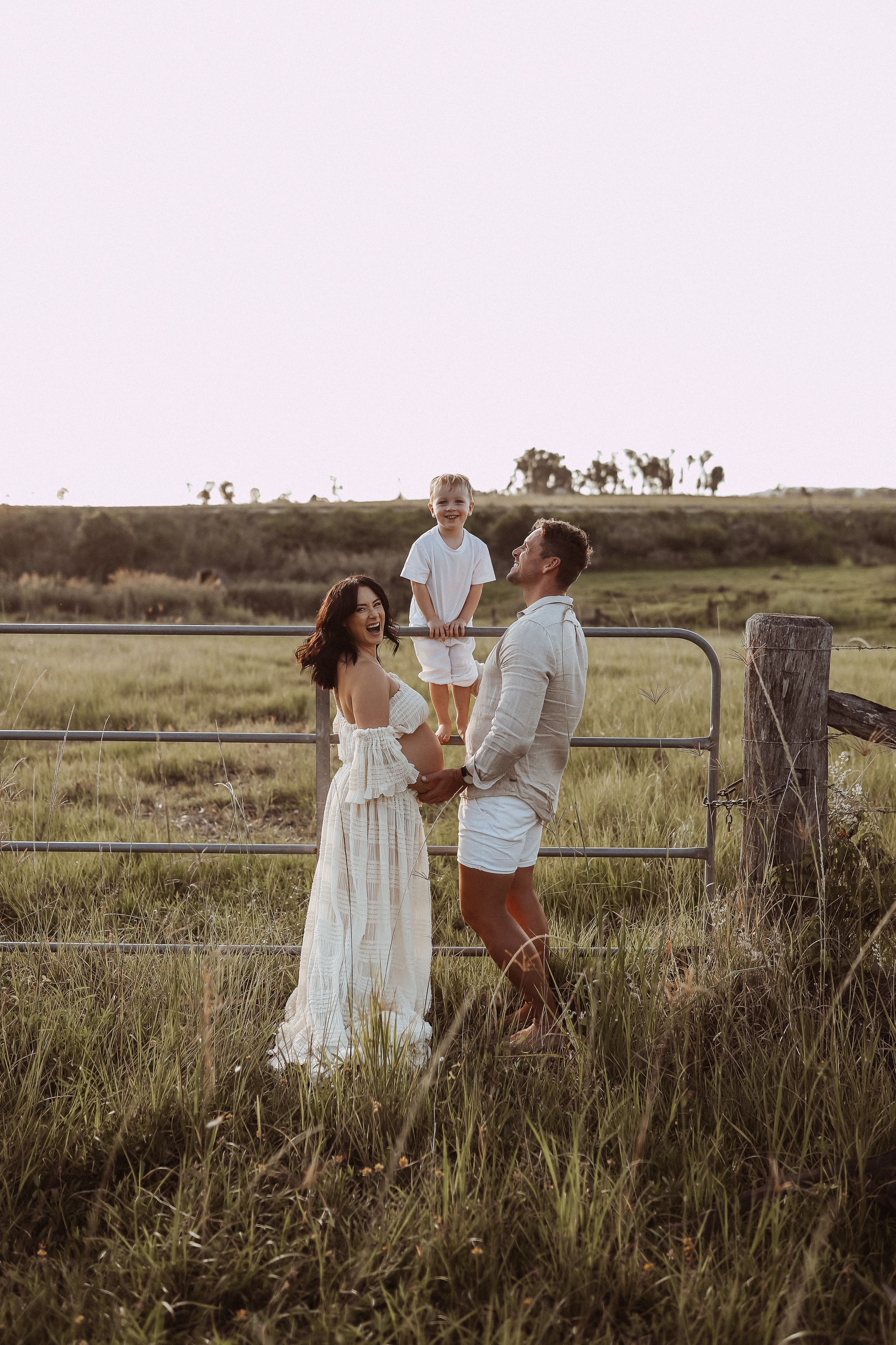 Family stands together near rustic fence in golden field at sunset.