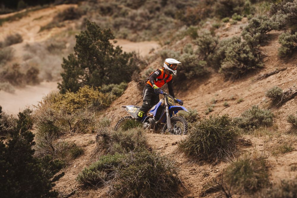 A motocross rider in white and orange gear navigates a steep sandy hillside on a blue dirt bike.