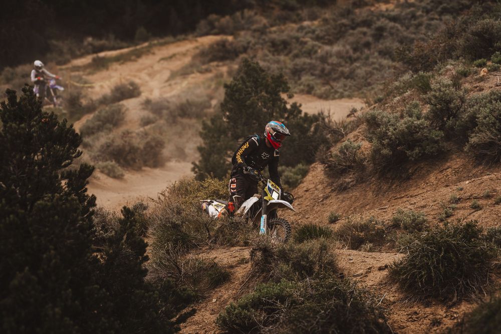 A motocross rider navigates a rugged dirt hillside trail surrounded by sparse shrubs during an off-road race.