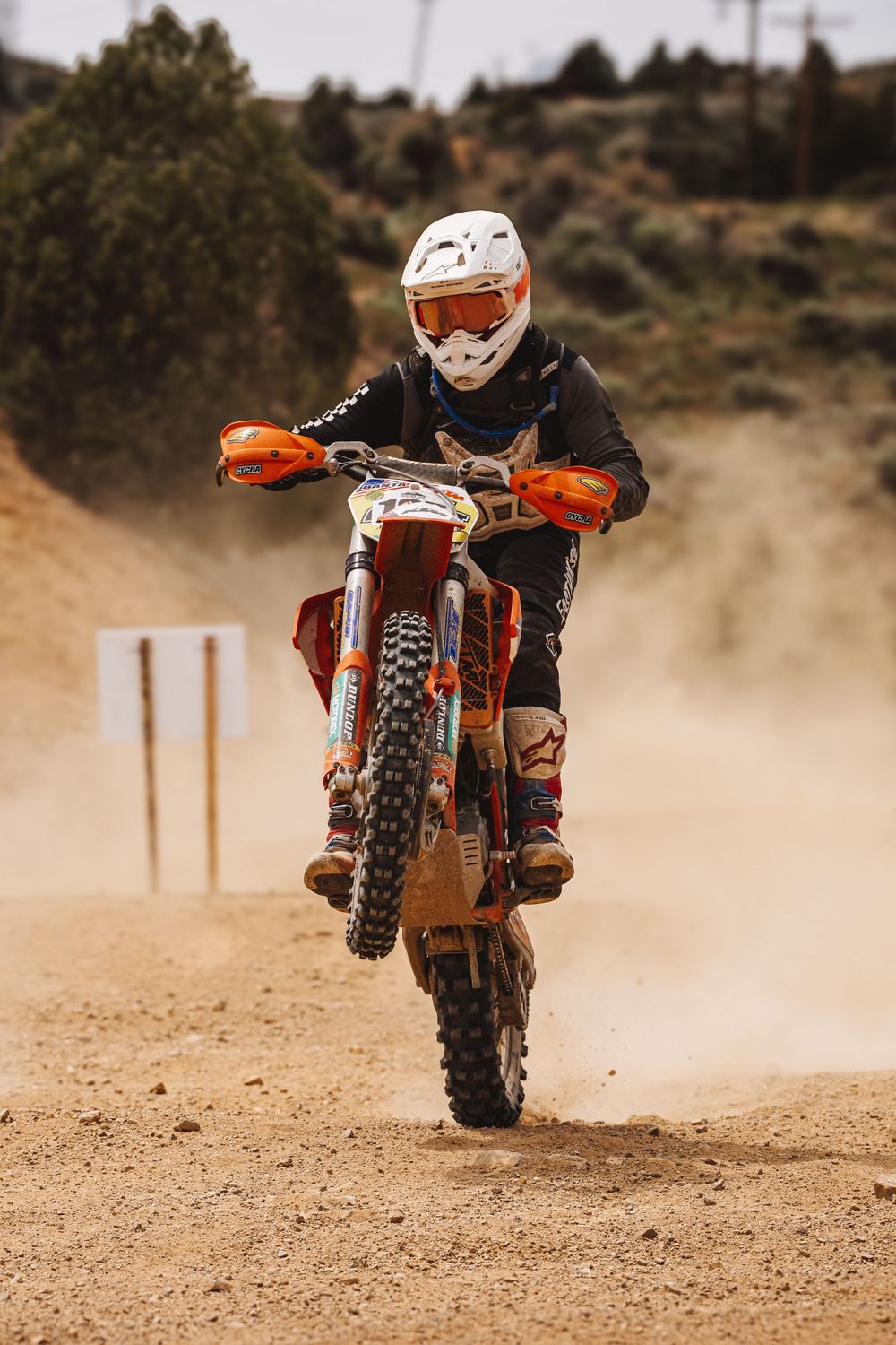 Motocross rider on orange KTM dirt bike racing through dusty off-road track wearing white helmet and black gear.