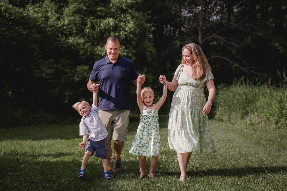 A family walks together and holds hands while enjoying a sunny summer day in a grassy field near the woods.