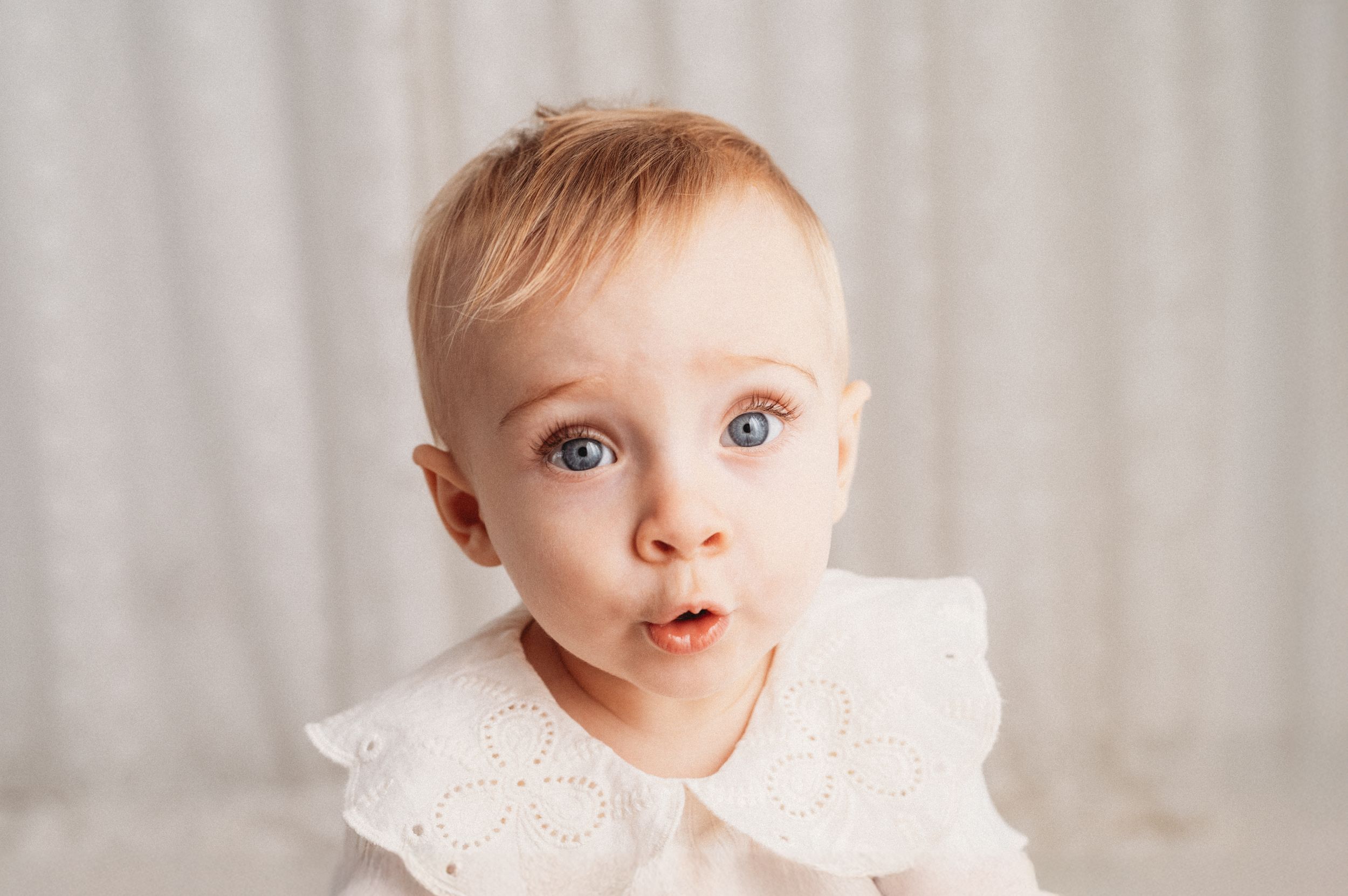 Baby wearing a white lace dress poses in a soft studio setting with natural lighting and neutral backdrop.