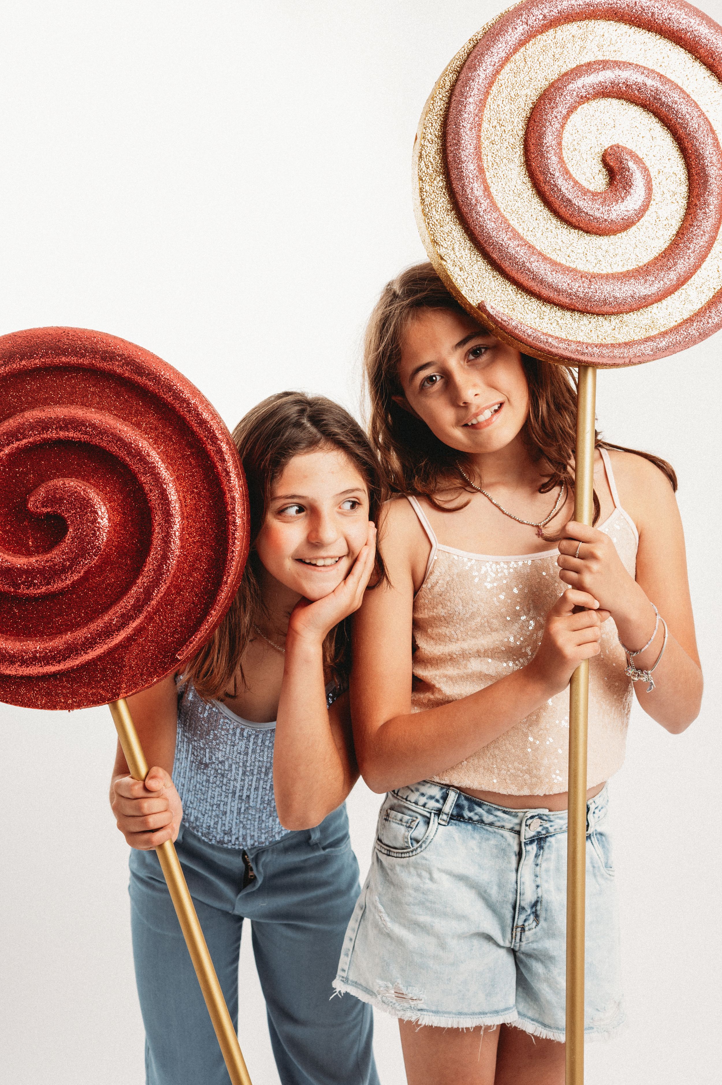 Two young people pose with oversized red and pink glittery lollipop decorations against a white background.