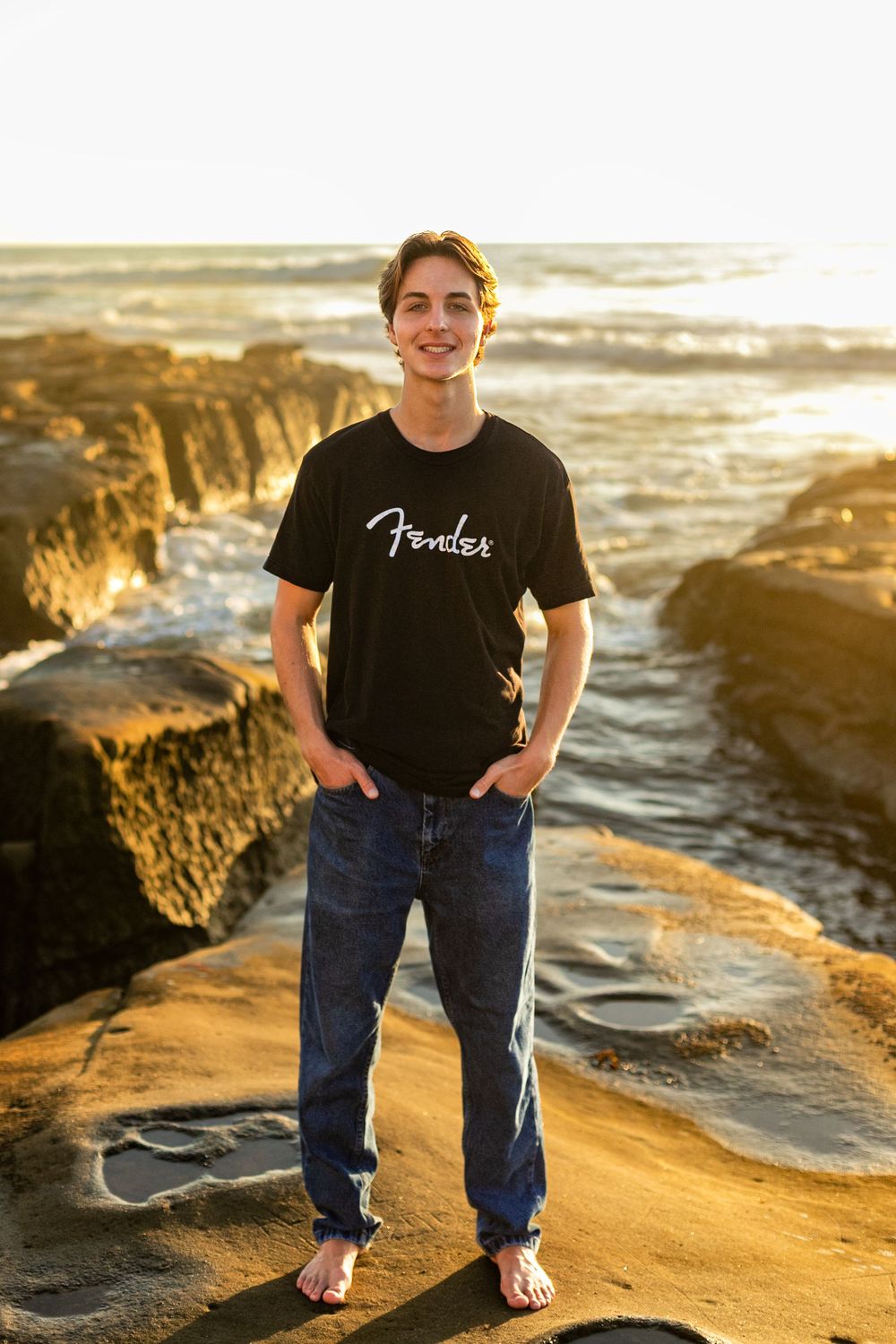 Person in black Fender t-shirt and jeans stands on golden rocky beach at sunset near ocean waves.