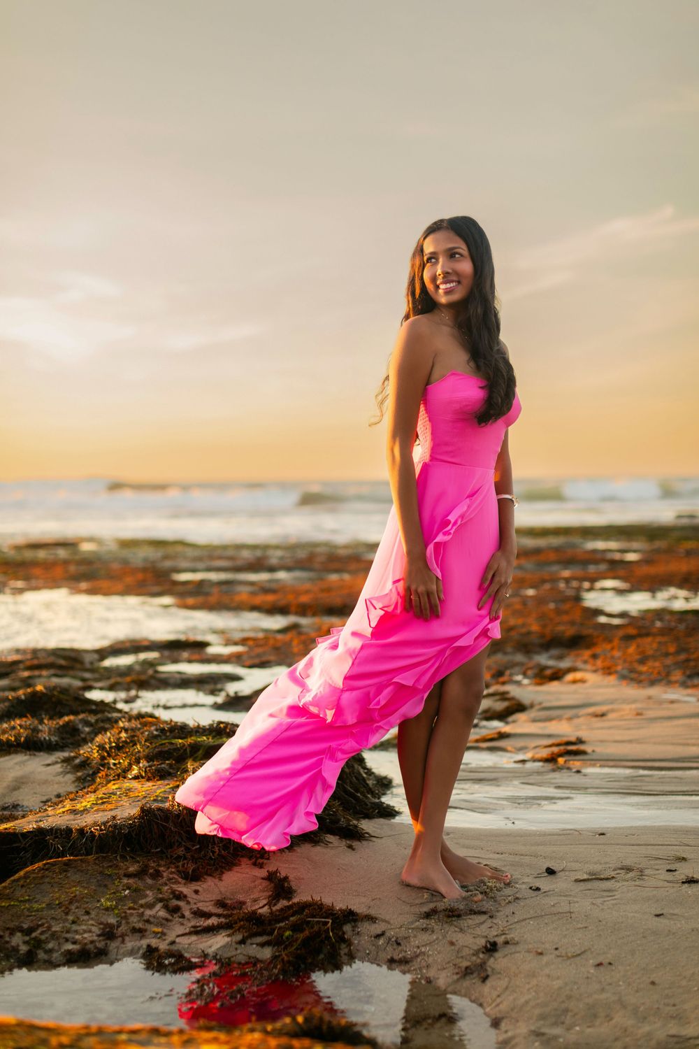 Woman in flowing pink gown poses barefoot on beach during golden sunset as waves crash against rocky shore.