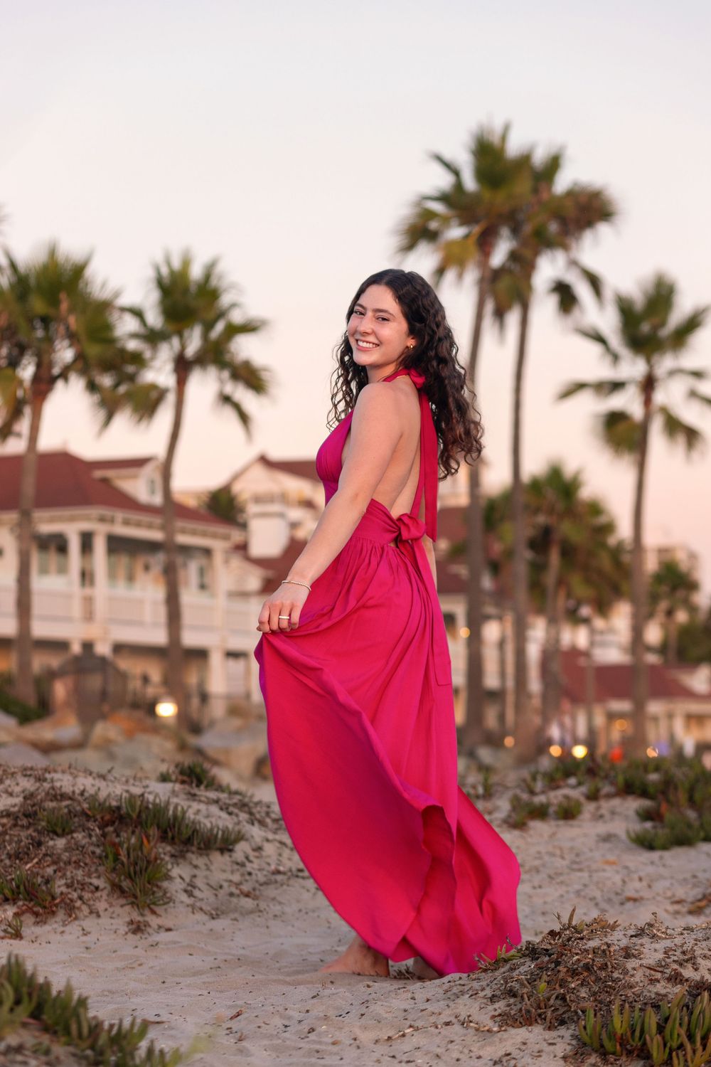 A flowing red dress catches the breeze as someone poses on a beach with palm trees and houses in the background at sunset.