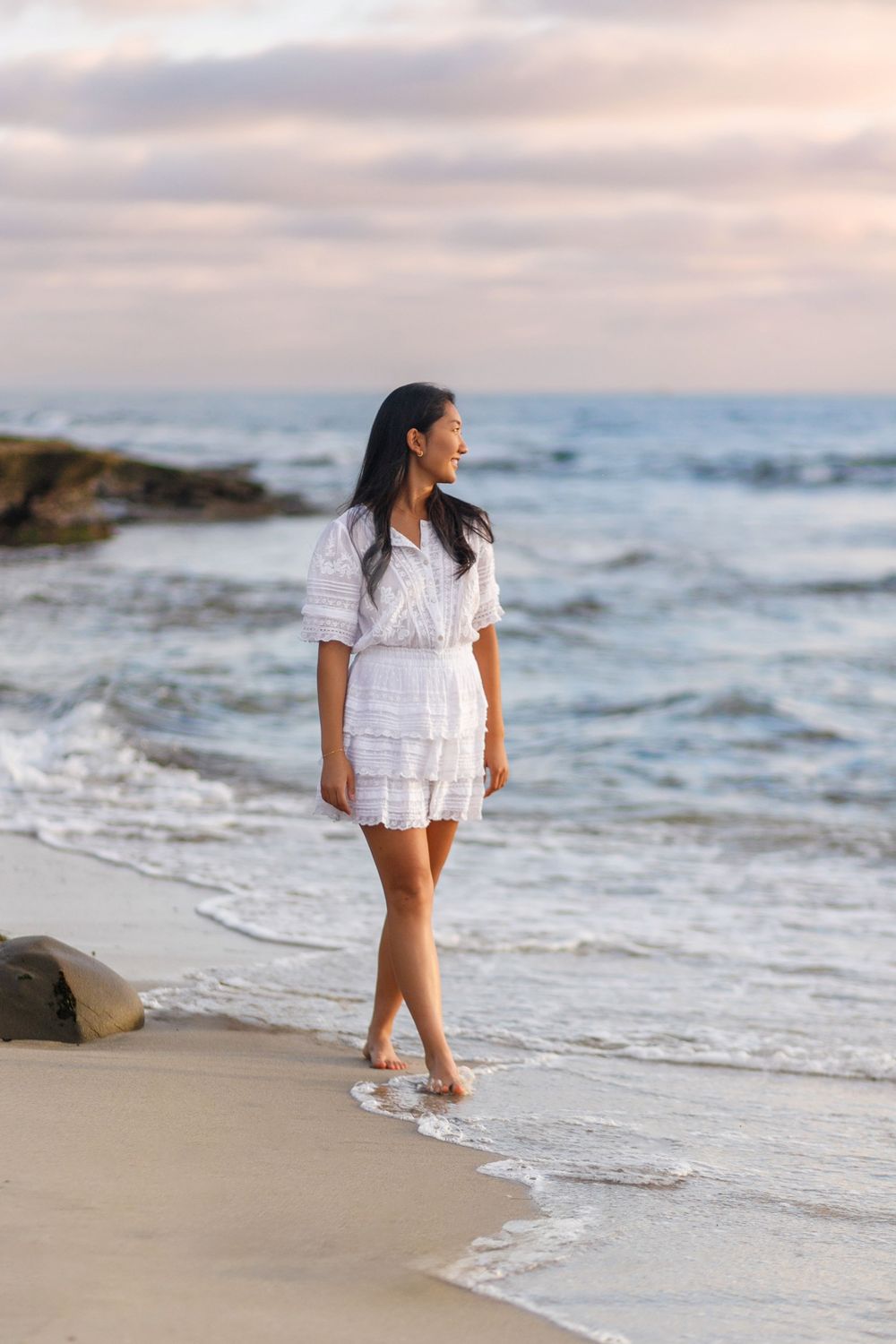 A person in a white dress walks along the shoreline as waves crash on a cloudy beach at sunset.