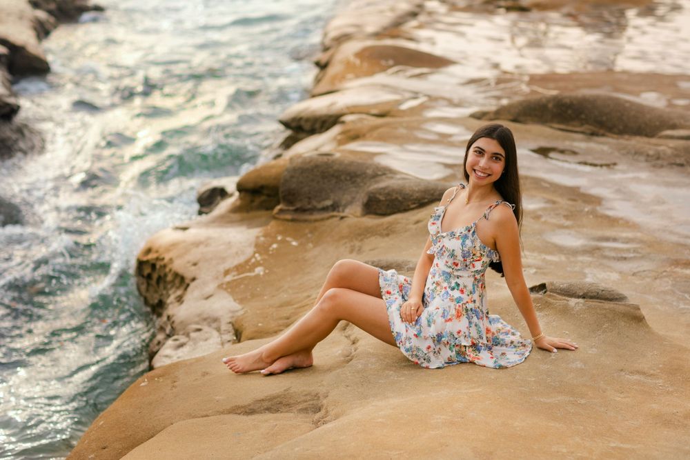 A summer dress clad figure sits gracefully on rocky coastal cliffs as ocean waves crash against the shore at sunset.