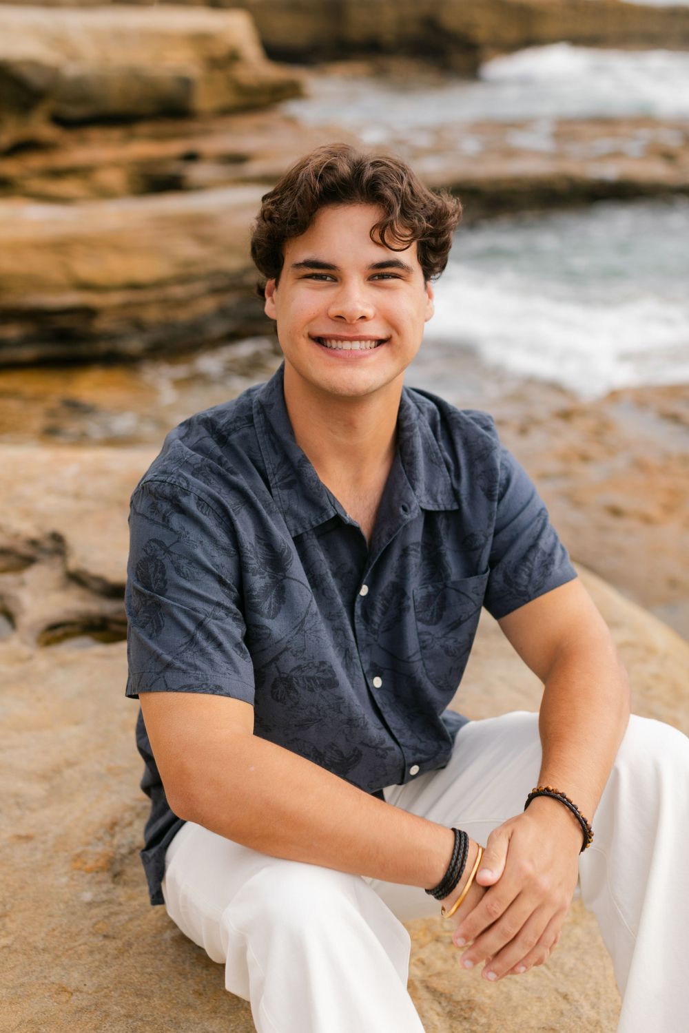 A young person in a navy button-down shirt and white pants sits smiling on coastal rocks near the ocean.