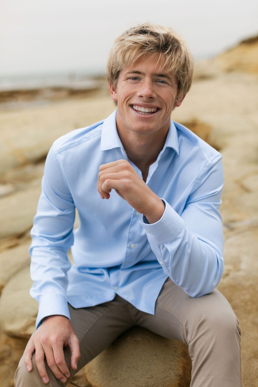 Person in light blue button-down shirt and khaki pants sitting outdoors and smiling on rocky terrain.