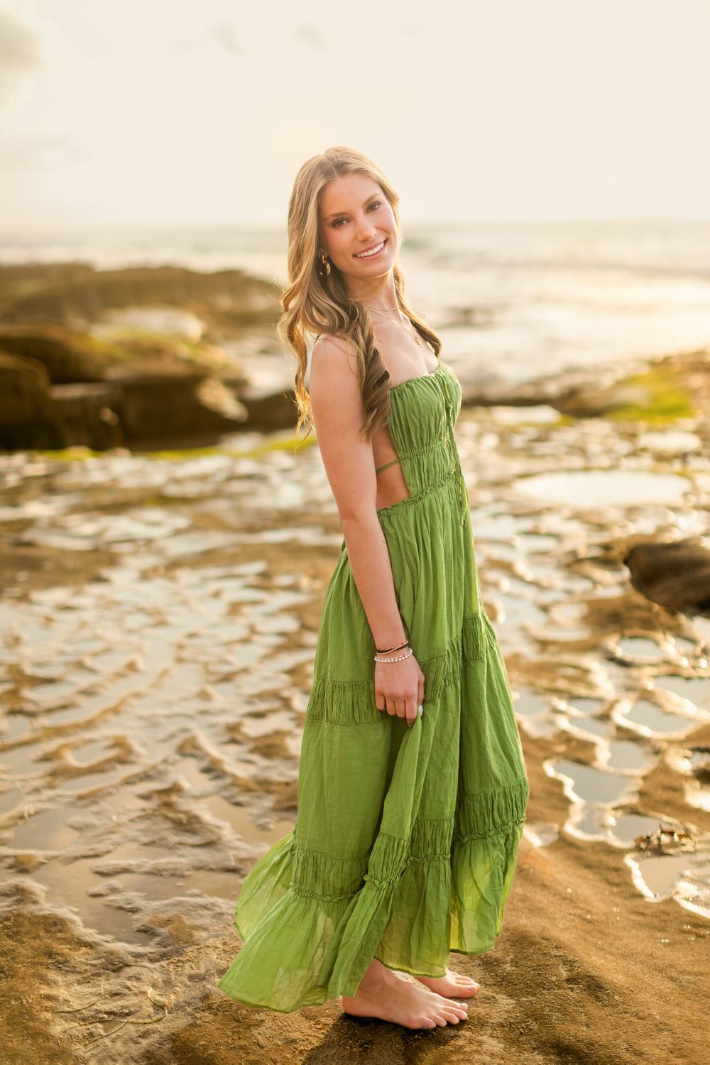 A figure in a flowing green maxi dress stands barefoot on rocky beach terrain during golden hour sunset.