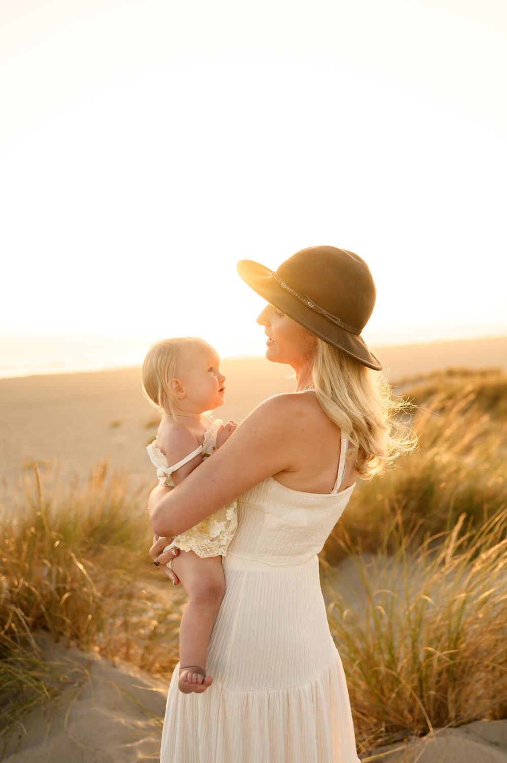 mommy and me photoshoot at Ventura beach on grassy sand dunes of mom in white dress singing to baby girl, by Santa Barbara photographer