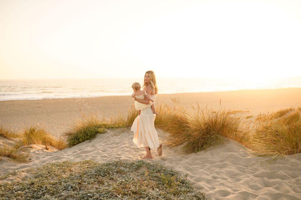 mommy and me photoshoot at Ventura beach on grassy sand dunes of mom in white dress walking with baby girl, by Santa Barbara photographer