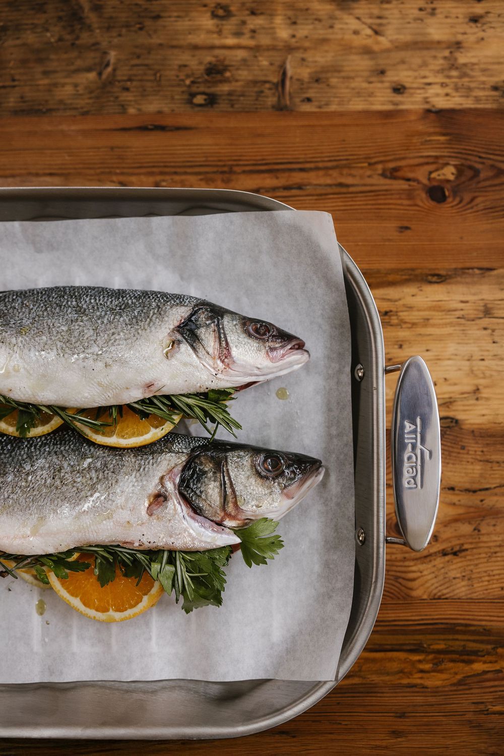 Two raw sea bass fish with lemon and herbs on parchment in a roasting pan.